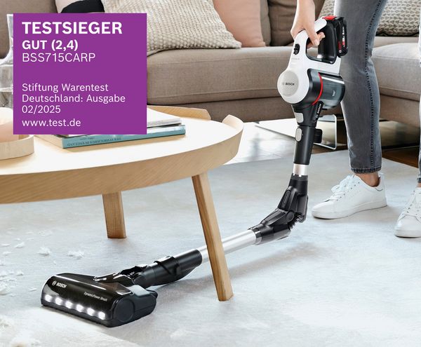 Eine Person verwendet einen Bosch Akkusauger auf einem Teppich unter einem Couchtisch, mit einem sichtbaren „Testsieger“-Abzeichen in der Ecke. 