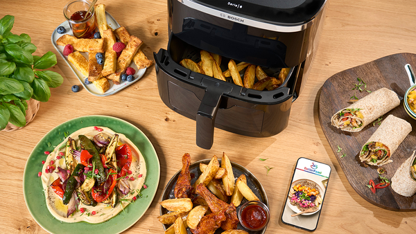 Der Bosch AirFryer auf einem hellen Holztische mit vielen Gerichten in Schalen angerichtet und dem Smartphone mit der BetterFood App.