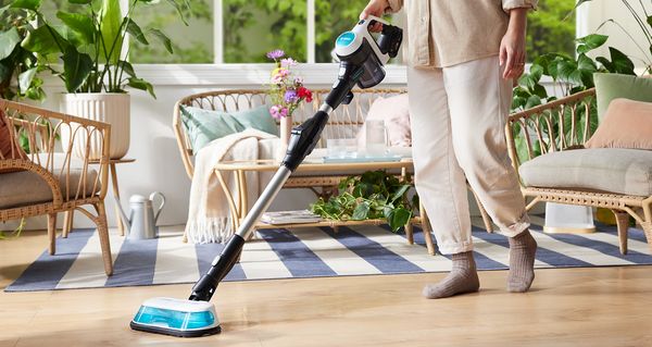 Eine Person reinigt mit dem Unlimited 7 ProHygienic Aqua Akku-Staubsauger von Bosch ihren Laminatboden. Im Hintergrund stehen Sofa und Sessel aus Ratan sowie mehrere Pflanzen.