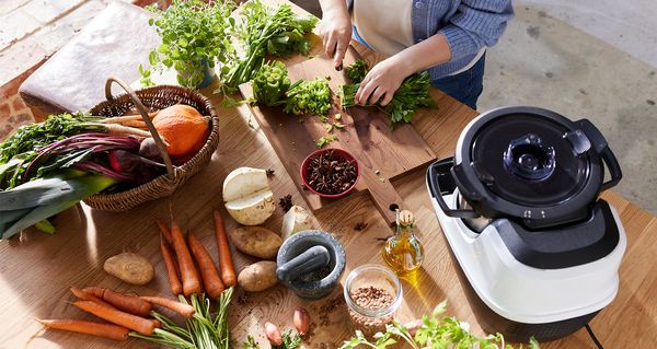 Eine Person steht an einer Arbeitsplatte und schneidet auf einem Holzbrett ein Bund Petersilie klein. Um das Holzbrett verteilt stehen ein Korb mit Herbstgemüse sowie der Cookit von Bosch. Dazwischen liegen Möhren, ein angeschnittener Rettich sowie ein Mörser und Gewürze.