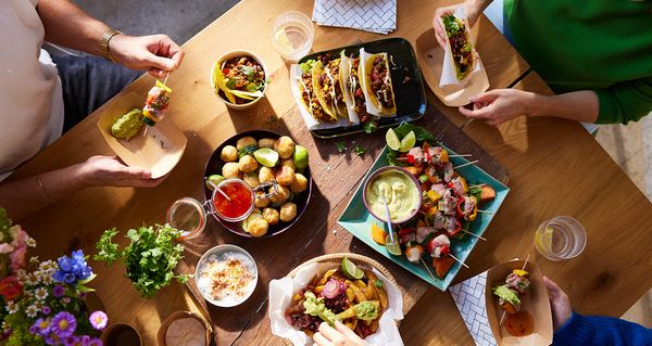 Drei Personen sitzen an einem Holztisch und genießen verschiedenes Fingerfood wie Tacos, Pommes mit Avocadodip und Fleisch-Gemüse-Spieße. Die Atmosphäre wirkt entspannt und einladend.