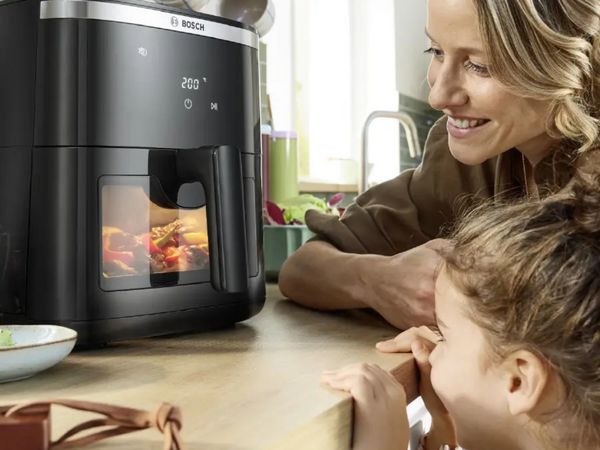 Madre e bambina guardano sorridenti il cibo in cottura nella friggitrice ad aria Bosch con finestra illuminata