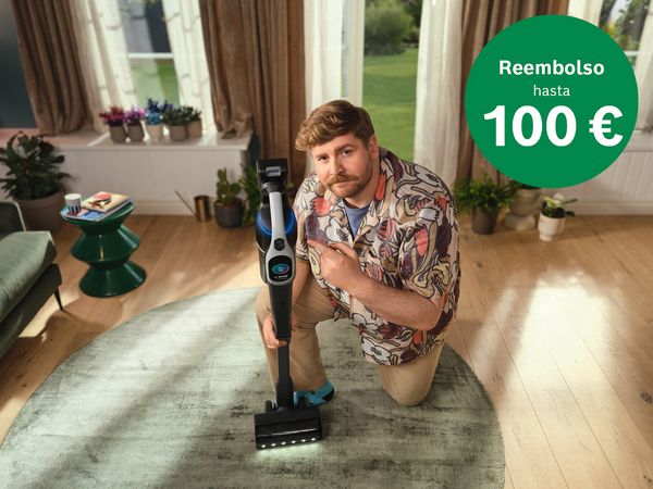Hombre de rodillas, señalando una aspiradora moderna en el suelo. Un círculo verde indica un reembolso de hasta 100 €.