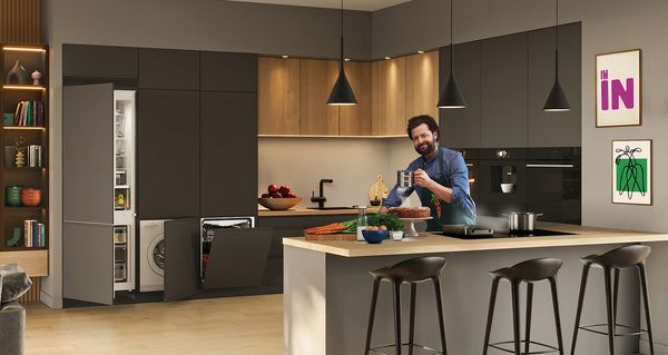 Hombre en el delantal verde de pie en la cocina, señalando a las verduras en la tabla de cortar.