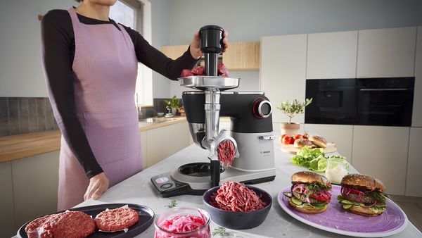 Person benutzt Bosch-Fleischwolf in der Küche und macht Burger, frische Zutaten stehen daneben.