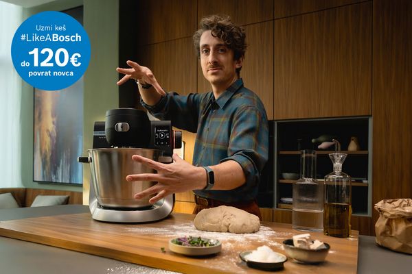Osoba demonstrira Bosch kuhinjski mikser na drvenoj radnoj ploči, okružena staklenim posuđem i kruhom