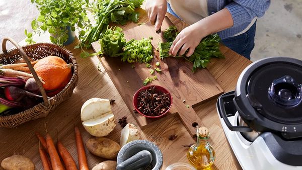 Eine Person steht an einer Arbeitsplatte und schneidet auf einem Holzbrett ein Bund Petersilie klein. Um das Holzbrett verteilt stehen ein Korb mit Herbstgemüse sowie der Cookit von Bosch. Dazwischen liegen Möhren, ein angeschnittener Rettich sowie ein Mörser und Gewürze.