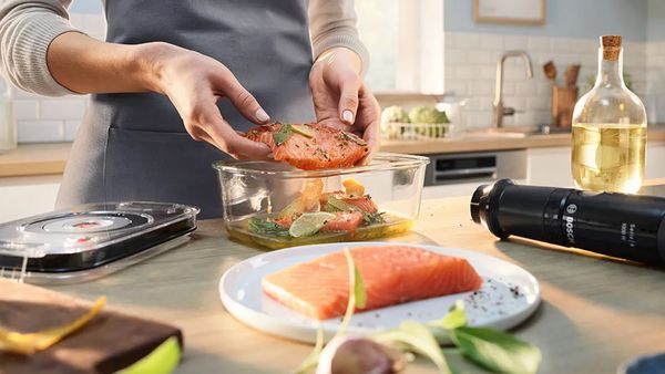 Eine Frau mit Schürze gibt ein mariniertes Fischfilet in einen Glasbehälter. Daneben liegen der Vakuumierdeckel und der ErgoMaster Serie 4 von Bosch mit Vakuumier-Funktion.