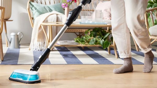 Eine Person reinigt mit dem Unlimited 7 ProHygienic Aqua Akku-Staubsauger von Bosch ihren Laminatboden. Im Hintergrund stehen Sofa und Sessel aus Ratan sowie mehrere Pflanzen.