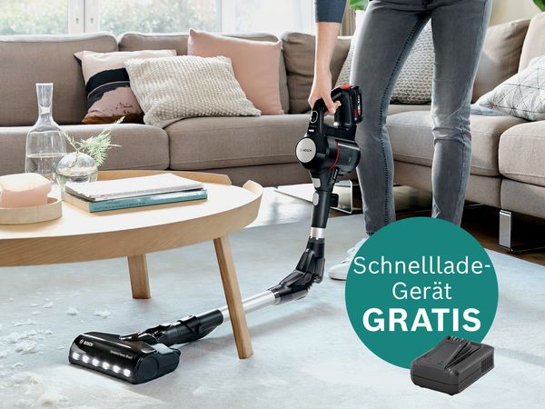 Eine Person verwendet einen kabellosen Staubsauger von Bosch auf einem mit Teppich ausgelegten Wohnzimmerboden neben einem Couchtisch und wirbt für das Angebot eines kostenlosen Schnellladegeräts.