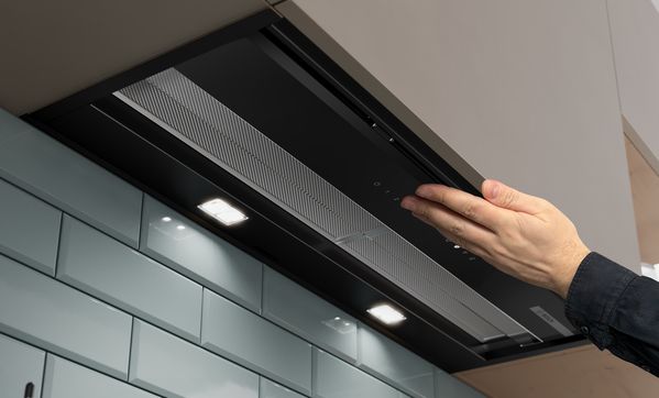 Изглед отдолу на аспиратор за пълно вграждане, като с ръка се почуква по сгънатото стъкло, за да се включи сензорната светлина.