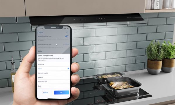Изглед отгоре на тенджери на плот, където се готви храна. По-горе, аспиратор за пълно вграждане извлича парата. Смартфон с екран на приложението Home Connect на преден план. 
