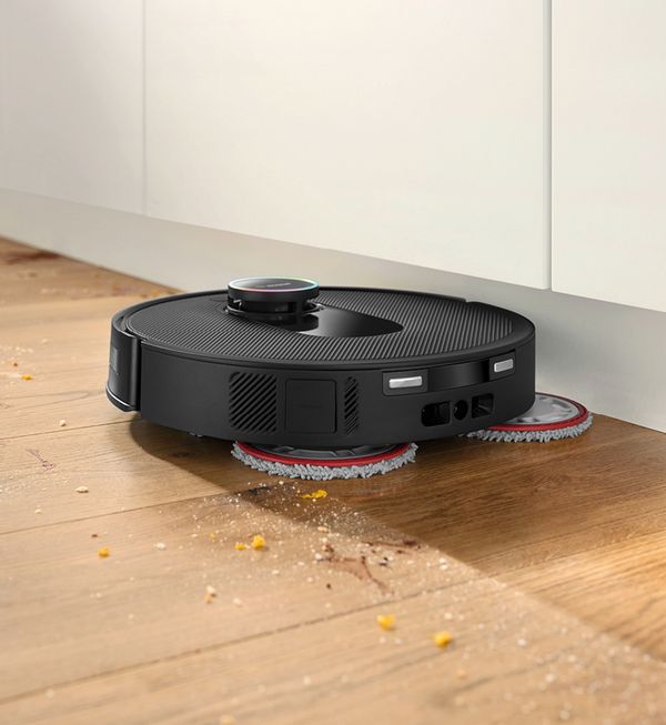 Ein schwarzer Saugroboter mit Wischtüchern reinigt den Boden unter einem Küchenschrank, umgeben von Krümeln.