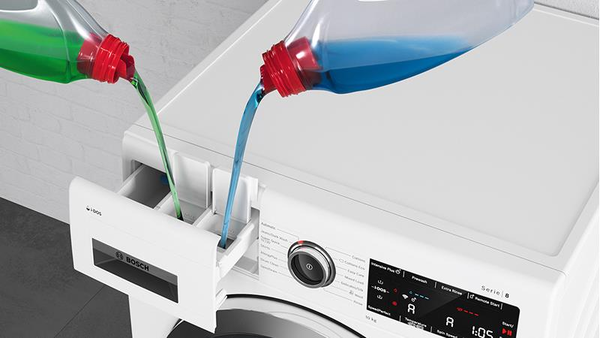 A bottle of blue liquid detergent is being poured into the detergent drawer of a white Bosch front-loading washing machine. 