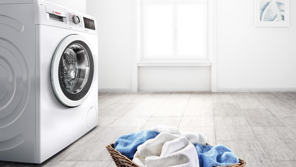 A close-up of a front-loading washing machine with its door open, revealing the drum, next to a wicker laundry basket filled with clean, folded towels, including white and blue ones, on a light-colored, possibly tiled, floor.  