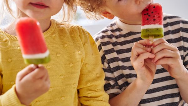 Es sind zwei Kinder zu sehen, ein Mädchen und ein Junge, die ein Stieleis in Wassermelonen-Optik essen.