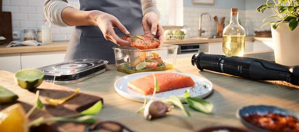 Eine Frau mit Schürze gibt ein mariniertes Fischfilet in einen Glasbehälter. Daneben liegen der Vakuumierdeckel und der ErgoMaster Serie 4 von Bosch mit Vakuumier-Funktion.