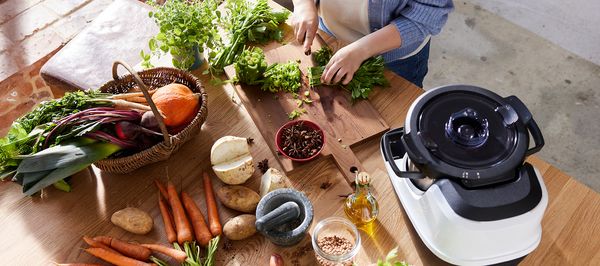 Eine Person steht an einer Arbeitsplatte und schneidet auf einem Holzbrett ein Bund Petersilie klein. Um das Holzbrett verteilt stehen ein Korb mit Herbstgemüse sowie der Cookit von Bosch. Dazwischen liegen Möhren, ein angeschnittener Rettich sowie ein Mörser und Gewürze.