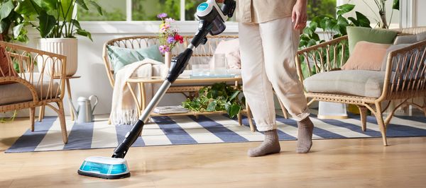 Eine Person reinigt mit dem Unlimited 7 ProHygienic Aqua Akku-Staubsauger von Bosch ihren Laminatboden. Im Hintergrund stehen Sofa und Sessel aus Ratan sowie mehrere Pflanzen.