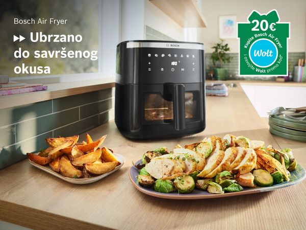 Friteza na vrući zrak na kuhinjskom elementu, ispred tanjuri s pripremljenom hranom, u kutu oznaka u obliku poklona s Wolt logom.
