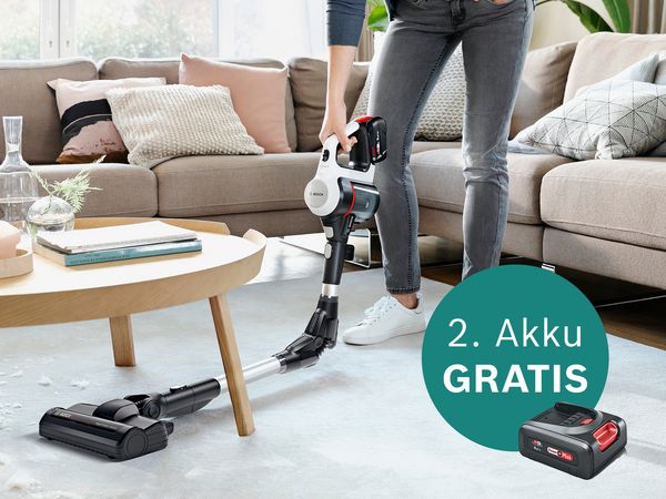 Eine Person saugt ein modernes Wohnzimmer mit einem kabellosen Bosch-Staubsauger und präsentiert dabei einen Werbeaufkleber für eine kostenlose zweite Batterie.
