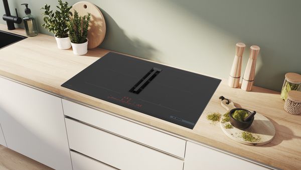 Zwart inductiekookplaat met afzuiging op houten keukentafel met potplant en andere keukenartikelen.