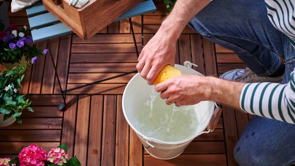 Eine Person kniet auf Terrassenfliesen in Holzoptik. Vor ihr steht ein weißer Wassereimer, über dem sie einen Schwamm auswringt. Im Hintergrund stehen ein Balkonstuhl und mehrere Pflanzentöpfe.