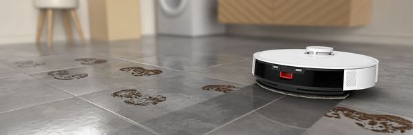 Un aspirapolvere robotizzato naviga su un pavimento piastrellato segnato da impronte fangose, evidenziandone la capacità di pulizia.