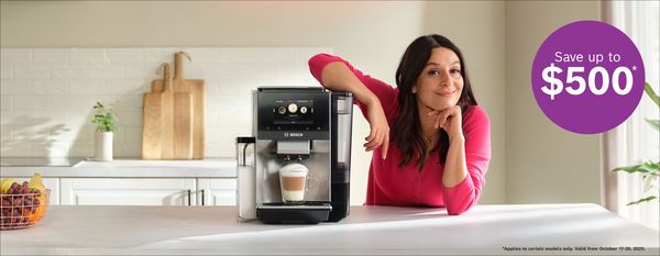 A woman in a pink sweater leans on a countertop, showcasing a Bosch coffee machine with a latte prepared. A promotional tag reads "Save up to $500.