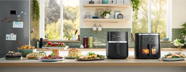 Two Bosch Air Fryer models positioned side by side on a bright kitchen counter. Several plates and bowls of food, including herb bread, chicken, roast, and fries, are placed next to the appliances, showcasing dishes made using the Air Fryers.
