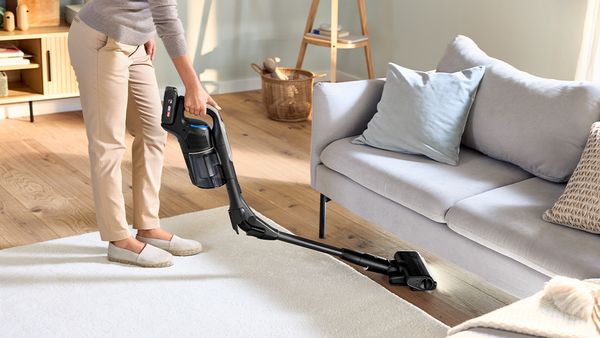 Eine Frau saugt mit einem Unlimited Staubsauger von Bosch mit knickbarem Rohr den Fußboden unter einem Sofa.