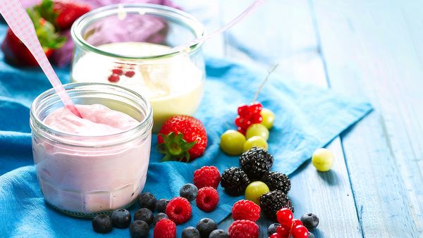 Два стъклени буркана с крем рикота, единият розов, а другият бял, стоят върху синя салфетка. Те са заобиколени от различни пресни плодове като малини, боровинки и ягоди.