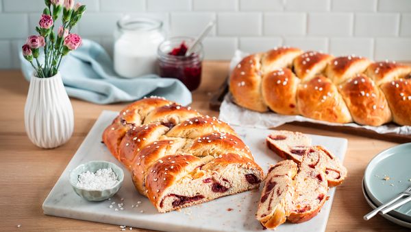 Два козунака с мая и перлена захар на дървена маса. Единият е нарязан върху мраморна дъска, разкривайки пълнеж от череши. На заден план се виждат буркан със сладко и цветя.