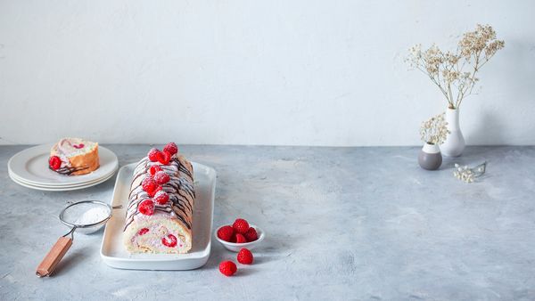 Руло с малинов крем върху бяла чиния, украсено с пресни малини, пудра захар и шоколадова глазура. На заден план е сервирано парче в чиния.