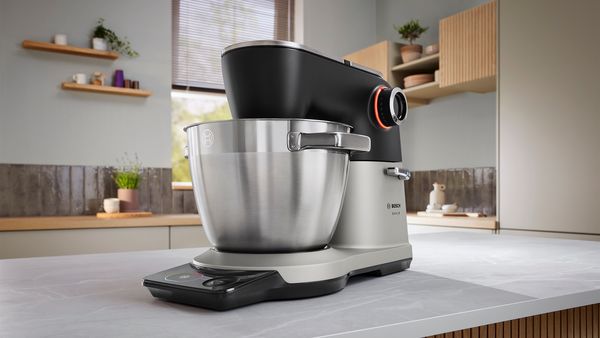 Върху модерен кухненски плот е поставена кухненска машина Bosch Серия 8 в черно и сребристо. Уредът е с голяма купа от неръждаема стомана и осветен контролен бутон.