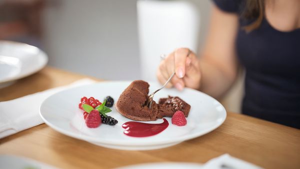 Ръка използва лъжица, за да отреше хапка шоколадова лава торта, от която изтича разтопен шоколад. Десертът е гарниран с пресни плодове, мента и пудра захар върху бяла чиния.