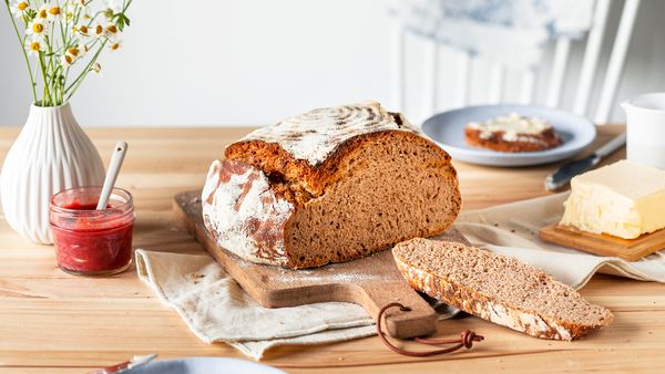 Поръсен с брашно рустик хляб с квас върху дървена дъска, с отрязана една филия. Масло, сладко и ваза с цветя на масата за уютна обстановка за закуска.