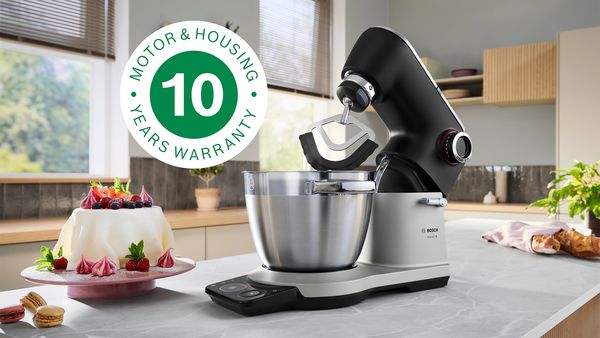 Кухненска машина Bosch Серия 8 в черно и сребристо на кухненски плот. Етикетът показва „10 години гаранция на мотора и корпуса“. Наблизо има украсена торта и макарони.