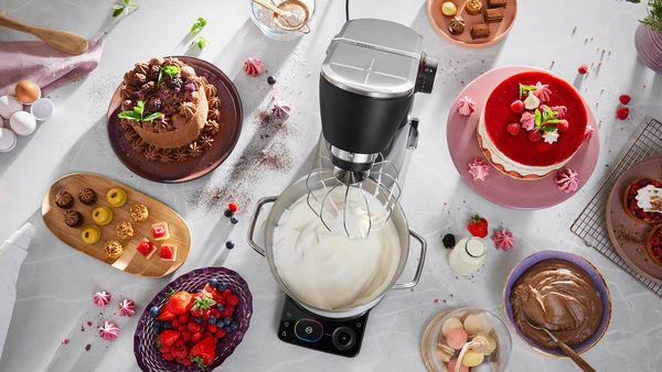 Кухненска машина на кухненски плот до гарнирана основа за пица с момче, дегустиращо съставки, което стои до мъж в кухнята. 