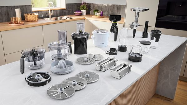 Комплект приставки за кухненска машина Bosch Серия 8, изложен на плот. Кухненска машина Bosch. До нея човек използва цитрус преса, блендер и шредер.