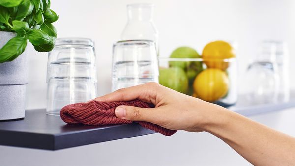 Es ist eine Hand zu sehen, die mit einem roten Staubtuch ein Regalbrett abwischt, auf dem mehrere Gläser, ein Topf Basilikum und eine Glasschale mit Zitrusfrüchten stehen. 
