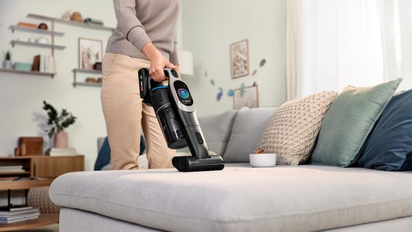 Eine Frau saugt mit dem Handteil eines kabellosen Staubsaugers inklusive Polsterdüse ihr hellgraues Sofa ab.