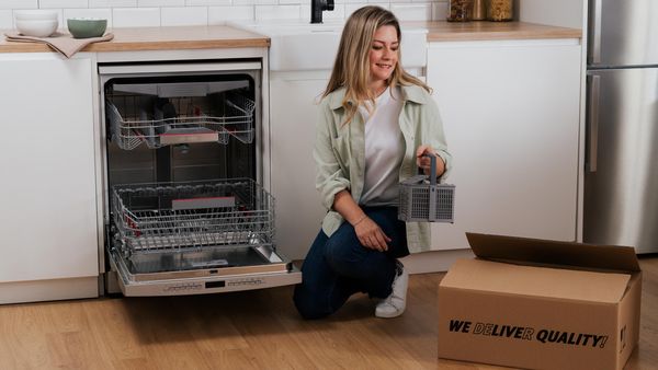 Una mujer se arrodilla junto a un lavavajillas abierto, sosteniendo una cesta para cubiertos y cerca de ella una caja de cartón con una etiqueta que dice "¡ENTREGAMOS CALIDAD!".