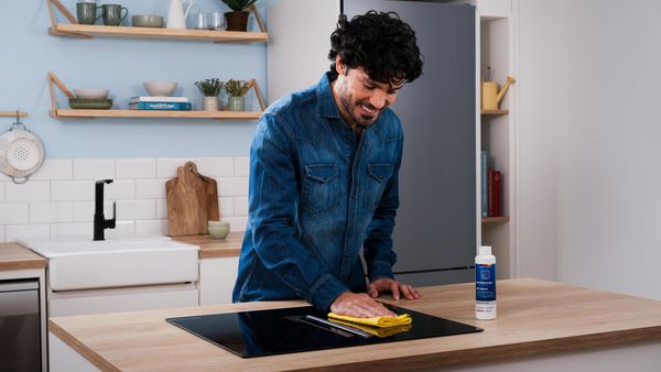 Una persona con una chaqueta vaquera limpia una placa de cocina negra con un paño amarillo, rodeada de una cocina moderna con estantes y plantas.