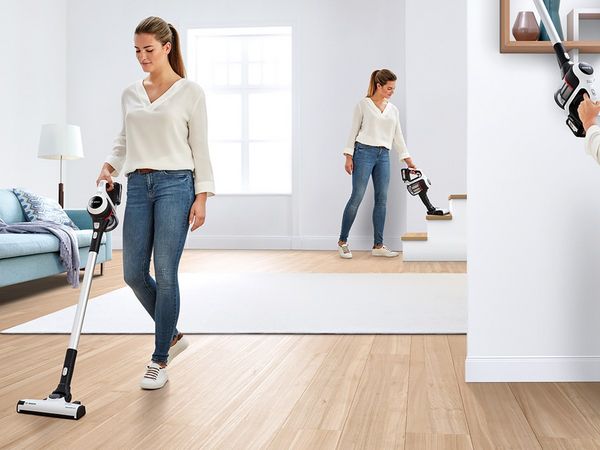A woman vacuums the carpet in a tidy living room, surrounded by furniture and natural light from a window.  