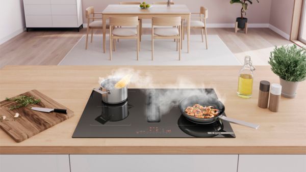 Модерна кухня с индукционен готварски плот, на който се готвят паста и скариди, с подправки и олио наблизо.