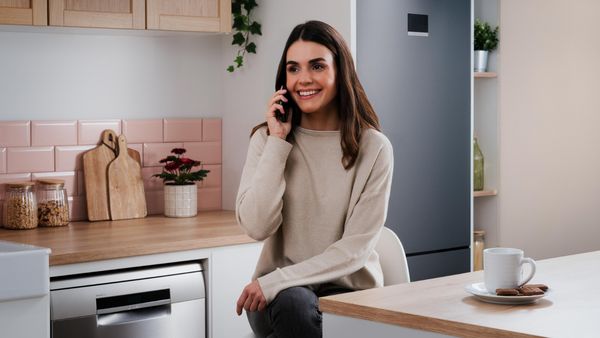 Una mujer en una acogedora cocina está sentada en un taburete, sosteniendo un teléfono, con una taza de té y galletas en la mesa a su lado.