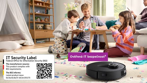 Rodina hraje u nízkého stolu v útulném obývacím pokoji, robotický vysavač s IT bezpečnostní samolepkou je v popředí.