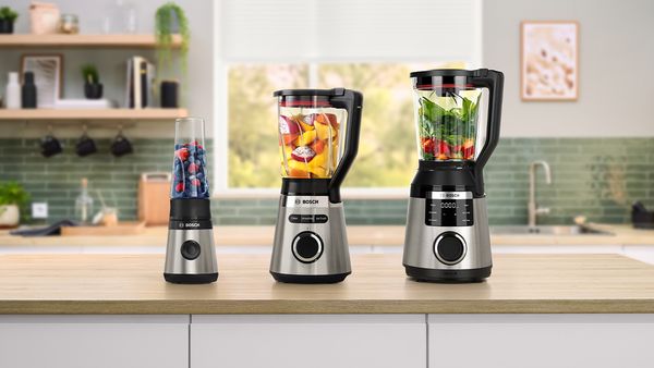Drei Bosch-Standmixer in Schwarz und Edelstahl stehen auf einer hellen Kücheninsel: links mit Beeren, mittig mit gelben und roten Früchten, rechts mit grünem Blattgemüse und roten Früchten gefüllt.