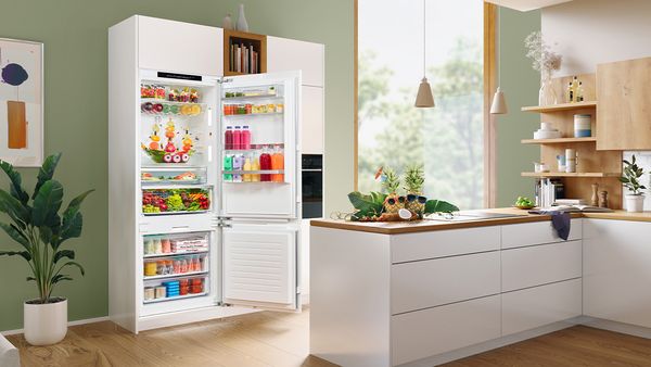 Blick in eine moderne, helle Wohnk&uuml;che: Links steht eine ge&ouml;ffnete, gut gef&uuml;llte Bosch-K&uuml;hl-Gefrier-Kombination, rechts davon eine L-f&ouml;rmige K&uuml;cheninsel mit Holzarbeitsplatte.